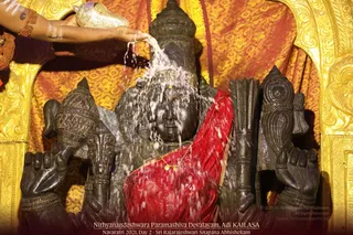 A black stone deity murti of a goddess is being bathed with water during abhishekam at Nithyanandeshwara Paramashiva Devalayam, Adi KAILASA. The murti wears a red silk drape edged in gold, a gold necklace and gold ornaments, and is framed by an ornate gold-embroidered backdrop. A hand at the upper left, adorned with red thread and a bangle, pours water from a golden vessel over the figure's head as water spills in a stream. Only the partially visible arm of a person is present; the murti is the focus. The photo is captioned Navaratri 2021, Day 2 - Sri Rajarajeshwari Snapana Abhishekam.