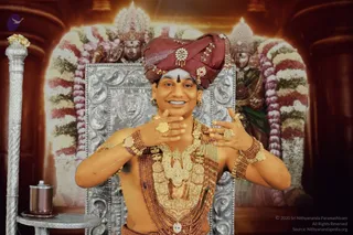 A smiling bare-chested man stands before a silver-framed throne backdrop, wearing a maroon turban with jeweled brooches, a tilak on his forehead, heavy gold necklaces, rudraksha malas, gold bangles and rings, and making hand gestures with both hands. Behind him, garlanded deity murtis of a royal divine couple with floral arches frame the scene, and a silver ritual stand with a copper vessel stands to the left.