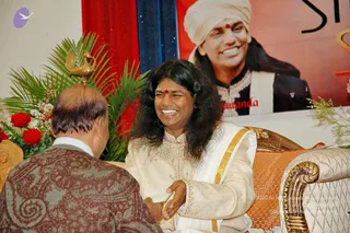 A photo of a seated man with long, dark, swept-back hair, smiling broadly, wearing a cream and gold embroidered kurta with a white and gold shawl and a red bindi on his forehead, seated on an ornate cream and gold armchair with an orange cushion. He clasps the hand of an older bald man with his back to the camera, whose brown paisley jacket and white collar are visible. Behind them a large banner shows a smiling man in a white turban and dark embroidered jacket with the word 'nanda' visible. Red roses, palm fronds and a golden bird ornament decorate the stage.