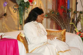 A photograph of a long-haired person in a white ceremonial outfit with gold embroidered trim sits on a white-draped sofa, head bowed and eyes closed, one hand resting on a gold brocade cushion. Behind them stand a tall glass vase of pink and white flowers and a fan of peacock feathers, with gold and red fabric drapes in the background. A pink cloth covers a small table at left, and a few pink and yellow flowers appear at right. The clothing and setting suggest a seated devotional or satsang setting. a person: the long swept-back hair, white ceremonial garments with gold trim and the seated central position match appearance cues for the page's subject, but the face is not clearly visible enough to confirm.