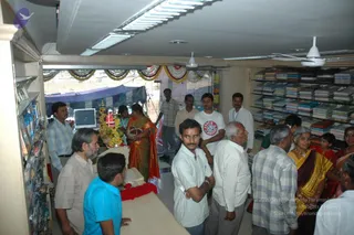 Interior of a bookshop or business premises during an inauguration. A woman in an orange and green sari stands near a table laden with fruit, flowers and a small decorated arrangement, with a red flower garland laid on the counter in front. Men and women in casual shirts, a white t-shirt and a checked shirt crowd around, some looking on. Shelves of books line the right wall and a computer monitor sits on the left counter. Marigold and mango-leaf festoons are strung across the open doorway, and a blue tent awning is visible outside.