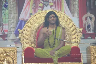 A man sits cross-legged on an ornate golden throne with red cushioned armrests, speaking into a microphone on a stand. He has long dark hair, a small red bindi on his forehead, a dark rudraksha mala around his neck and a light green shawl draped over his bare chest and legs. His right hand is raised near his chest in a gesture of teaching. Behind him a large backdrop shows a painted figure in pink and blue robes with a garland, and a decorated shrine with a small deity murti and white flowers appears at the right edge. The setting suggests a satsang discourse. Appearance-based label: Swamiji (probable), seated on the throne with long hair, rudraksha mala and green shawl.