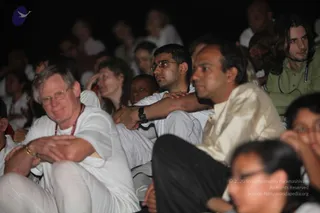 A dim evening satsang audience seated closely together on the ground, watching toward an unseen stage. In the foreground an older man with grey hair and glasses wears white clothes and a red lanyard, hands clasped. Beside him a young man in white and glasses rests his arms on a companion in a cream kurta. A woman with long hair and a young girl with glasses appear at the lower right, and many other attendees in white fill the background.