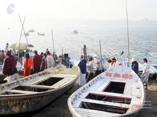 Photograph of a riverbank landing at the Triveni Sangam in Allahabad, with several wooden ferry boats drawn up on sand. One boat has the painted name "Bank of Baroda" on its side. A group of people in mostly white and red-brown clothing, including a woman in orange and a man in an orange-and-black knit cap, stands and boards near the boats. Long bamboo poles stand upright among the boats, and more boats and birds are visible on the hazy river behind.