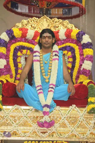 A man with long dark hair and a red bindi on his forehead sits cross-legged on a gold ornate throne covered with garlands of white jasmine, yellow and pink marigolds and purple flowers. He wears a sky-blue dhoti and a matching shawl draped over one shoulder, a heavy gold bead mala and a gold chain. A red velvet cushion rests behind him beneath a red and gold canopy with tassels. A gold-carved base is decorated with white flower chains.