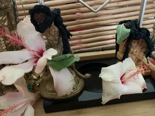Close photo of a small altar display on a black lacquered tray set against a woven bamboo backdrop. A brass deity murti sits in the centre, partly covered by a dark crocheted hair-like cloth and a green leaf, with a pale pink hibiscus flower in front. A second carved wooden figure on the right wears a similar dark crocheted covering, a green leaf and another hibiscus blossom at its base. No people are visible.
