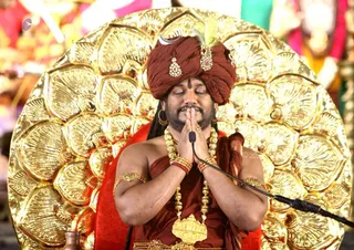 A man seated before a large golden fan-shaped backdrop with eyes closed and palms pressed together in namaste at a microphone stand. He wears a maroon-red turban with a crescent ornament, a feather and jewelled brooch, long gold necklaces, a large gold pendant, and maroon-red cloth draped over one shoulder with gold armlets. A small kalasha-like brass vessel is visible at the lower left. Behind him, blurred figures in pink and green clothing stand in the background. a person: the red turban, gold ornaments and ceremonial setting match the page's description of a satsang.