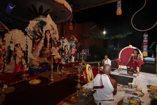Night-time photo of a temple-style puja set up outdoors beneath a canopy. On the left, a large white ornate arch frames a tall deity murti with multiple arms, holding weapons, with a lion figure at its base and other smaller murtis behind. A seated white-robed priest with shaved head faces the altar, and a second person in white sits near brass oil lamps (lamps) with flowers, fruit plates and offerings on red cloths. On the right, a figure in a red-and-gold ornate throne (peetha) wears a red sari, gold necklace and bangles. Strings of marigold garlands, hanging decorative banners and a green leaf festoon decorate the scene.