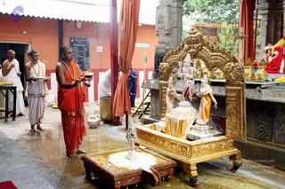 A temple abhishekam scene in an orange-walled hall. A priest in an orange-saffron dhoti and a shawl, wearing a rudraksha-style mala and gold-accented neckwear, stands holding a lit oil lamp (aarti) toward a gilded shrine. Two other priests in white dhotis with gold borders stand behind him, one holding a plate of offerings. The shrine holds a white-anointed deity murti with attendant figures in gold garments, set in an ornate gold arch with a gold pedestal; water and flower petals wet the floor. A printed board on the orange wall lists items, and a red-clothed deity is visible at the right. Three to four priests are visible, not a crowd.