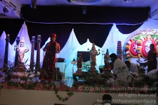 A photograph of a stage set for a ritual or festival performance, with a white draped backdrop shaped like mountain peaks and a dark curtain above. On the left stands a large silver-toned deity murti with raised hands. In the centre, a man in a red-brown dhoti and a matching orange-red turban wears a long garland of red flowers and a rudraksha-style mala, standing beside a smaller decorated murti. Further right are several more silver and gold deity figures, a kalasha-like pillar, and a floral arch with pink, green and yellow garlands framing a figure. Green leaves and vines line the platform's front edge. At the right foreground, men in white clothes sit cross-legged near the stage. Lights and a few audience silhouettes appear at the bottom. Only the man in red and the seated men in white are physical people; the statues are murtis.