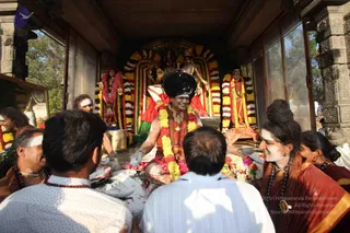 A photograph inside an open temple shrine with carved stone pillars and pale pillared walls. At the centre, a man with a large black hair bun and white forehead markings sits in a red and saffron robe, wearing a yellow and white flower garland and a gold necklace, smiling as he handles food and flowers on a tray. Around him, shrine statues are draped with marigold and yellow flower garlands, and a deity murti is visible behind. Several devotees with white-painted foreheads and dark rudraksha malas face him from the front, including a woman in a red sari on the right. A brass vessel sits near the centre of the shrine. The setting suggests a puja or padapuja ritual with offerings.