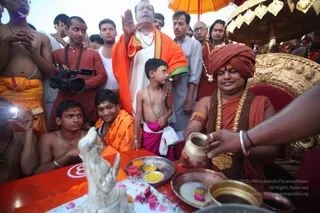 A photograph of a ritual at a riverside Kumbh Mela setting. A man with a crimson turban, orange robe, gold chest garland and rudraksha-style beaded necklace sits on an ornate golden throne-like seat under a golden umbrella, pouring liquid from a small brass pot into a metal plate of offerings. Nearby, a young boy in a pink dhoti with a long beaded necklace stands beside a seated man with an orange shawl, and a white stone deity murti sits on a red table. Several shirtless and orange-robed devotees with forehead markings crowd around, and a man in white holds an orange cloth. A cameraman stands at left. Pink flower petals and small bowls of coloured powder lie on the table.