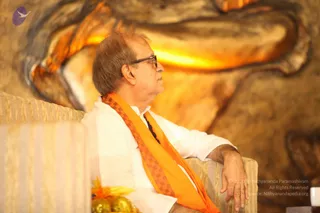 A seated man in profile wears a white collared shirt, black-rimmed glasses and an orange shawl with a dark checkered border draped over his shoulders. He rests one hand on the cushioned armrest of a pale gold upholstered chair. Behind him is a large warm-lit sculpted panel with glowing orange-gold carved forms. In the lower left corner, a bowl of yellow-and-gold wrapped sweets sits on a striped cushion. The scene is a quiet indoor setting with no other people visible.