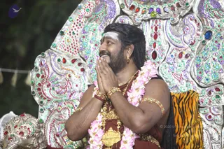 A seated man with long dark hair, a white vertical tilak on his forehead, and a warm smile sits in front of an ornate silver throne decorated with red, green, purple and blue jewel-like inlays. He wears a dark maroon-red garment, a large pink-and-white floral garland, gold necklaces, gold armlets and bangles, and a gold earring. His palms are pressed together in namaskar as he looks up and to the side. A tiger-striped orange cushion rests behind him and a decorated silver lion or elephant figure appears at lower left. Only one physical person is visible. Swamiji (probable): the long swept-back hair, ornate gold jewellery, garland and jewelled throne are the cues.