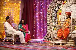 A stage satsang with three seated people before a golden temple-style backdrop and a draped curtain strung with fairy lights. On the right, a man in an orange-saffron robe, orange turban, and layered garland sits on an ornate silver throne with a leopard-print cushion, hands folded and smiling. Facing him on the left, a man in white kurta with a saffron shawl sits in a silver chair, and a woman in a green and pink sari holds a microphone and papers. Brass lamps and a decorated embroidered backdrop frame the scene. A watermarked caption at the bottom reads 'a person' context not printed; the central figure wears distinctive saffron robes, turban and garlands, matching the page's Ask The Avatar satsang.
