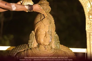 A brown-toned statue bust of a deity with a tall crown is being anointed with liquid poured from a golden vessel held by a bare arm wearing rudraksha beads, with the liquid streaming down the face and chest. The murti has ornate jewelled necklaces and shoulder pieces, set against dark night foliage and a gold-coloured arch at right. Caption text reads Nithyanandeshwara Paramashiva Devalayam, Adi KAILASA, Navaratri 2021, Day 7 - Abhishekam to SPH Nithyananda Paramashivam's Mula Vigraha.