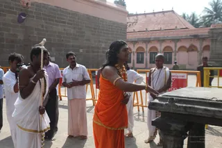 Outdoor temple courtyard beside a dark stone wall and pink-washed shrine building with arched colonnades. In the centre, a long-haired man in an orange saffron dhoti and shawl, wearing a gold-coloured ceremonial necklace, gold armlets and bangles, gestures with his right hand toward a dark carved stone platform. Around him, several men in white dhotis and shirts stand with hands clasped; one at left in white holds a tall staff topped with a bird finial, and a bald man in white with a sacred thread stands at right. Bright sunlight, yellow barriers and a red banner are in view.
