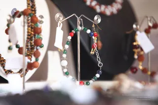 Close-up photo of jewellery displayed on silver hoop stands against a dark background. The foreground hoop carries a beaded bracelet or earring with white, red, green and dark blue beads, silver spacer rondelles and a small chain with a ring. Beside it, a hoop with rudraksha-style brown seed beads, tiger-eye style beads, green beads and gold accents, and a second hoop with red and brown beads hang on stands. Small paper price tags are attached. The background is blurred with a white bead necklace visible at the top. No people are visible.