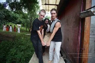 Outdoor photograph beside a wooden-and-tin shed wall, with lush green trees and bushes behind. Three men carry a long pale stone slab on their shoulders along a narrow concrete ledge. The man in front on the right has long swept-back hair, a red tilak on his forehead, a grey sleeveless wrap over a black top and light cream trousers, barefoot. The man on the left wears a black T-shirt with orange-red printed lettering and dark trousers. A bearded man in a white dhoti and black shirt helps in the middle. In the background, several people in white and red-orange traditional clothing stand on a grassy path. The photo credit overlay is visible at lower right.