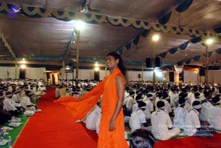 A photograph inside a large white tented hall at an evening convention. A man with long dark hair, wearing a flowing saffron-orange robe with a shawl draped over one arm, stands on a red carpet and smiles while turning toward the seated audience. Rows of participants in white clothes and white headbands sit cross-legged on mats on both sides of the carpet. Overhead lights, white tent drapery with blue and gold scalloped trim, and speaker boxes on poles frame the scene. Given the saffron robe, long hair and the page's note that Swamiji attended, the central figure is a probable Swamiji candidate.