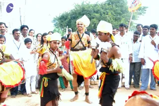 Outdoor photo of a folk-style performance on a dirt ground. Several male dancers wear yellow and gold turbans, bare upper bodies or gold-trimmed vests, black waist cloths with red and green sashes, and white forehead and arm markings. They hold metal swords and wave yellow fringed drum-like pieces. A crowd of spectators in white shirts, dhotis and casual clothes stands behind them under trees, with a saffron-and-white flag on a pole at the upper right. A small purple logo appears at the top left corner.