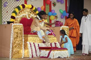 Photograph at a children's day event in a decorated hall. A man with long dark hair seated on an ornate gold-and-red throne wears a gold-embroidered tan robe and a multicoloured flower garland, and hands gifts wrapped in purple paper toward a young girl in a light blue dress who kneels before the throne. Thick white and maroon floral garlands cover the throne's base and the steps. Behind, a man in a saffron-orange robe with a maroon shawl stands beside a second man in white ritual clothing with a pendant necklace. Balloons in purple, blue, green and orange and paper decorations hang on a checked backdrop.