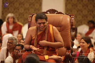 A smiling man with a bindi and an orange-saffron cloth draped over one shoulder sits on an ornate carved wooden throne with brown leather cushions, a cobra-head carving on the armrest. He wears heavy beaded gold-toned necklaces and a small microphone at his mouth, gesturing with an open hand. Around him, seated devotees in orange and red shawls and white clothes smile and listen in a hall with gold patterned wall panels. Appearance-based candidate: Swamiji (probable), seated on the throne with long swept-back dark hair, saffron cloth, and gold beaded necklaces.