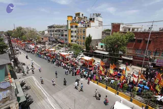 Aerial view of a long urban street procession in an Indian town on a bright day. Many people line both sides of the road and walk along it, some in orange and red clothing. Several decorated vehicles or carts with canopies, red and white awnings and orange banners move through the procession. Orange, yellow and red flags flutter from poles along the pavement, and shops, multi-storey buildings and parked cars line the street.