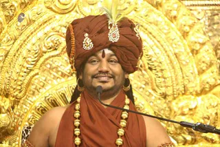 A man in a rust-orange turban with a gold-and-jewel brooch and a peacock-feather ornament sits before a microphone stand in front of ornate gold repoussé carving. He wears a matching rust-orange shawl, a rudraksha-style strand on the turban, and heavy gold beaded necklaces; a red bindi and white marking are on his forehead. He smiles toward the audience during a discourse. Swamiji (probable): the turban, saffron-orange shawl, ornate gold jewellery and rudraksha-style strand are the appearance cues.