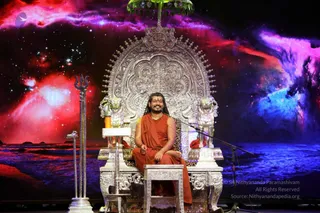 A man sits on an ornate silver throne with lion-head armrests and a silver canopy, on a stage before a cosmic backdrop of red, pink and blue nebulae. He wears an orange-saffron robe draped over one shoulder, long dark hair, and a rudraksha mala in his hands. A silver trishul stands at the left, a microphone stands at the right, and a silver stool sits in front of the throne. The appearance-based label Swamiji (probable) applies to this seated figure in saffron robes with long hair and a rudraksha mala, presented on a throne in an event setting.