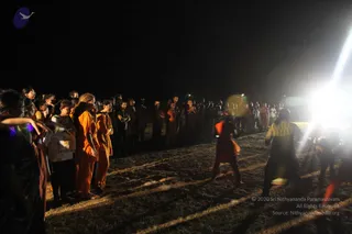 A night-time outdoor gathering on a dry open field. A large crowd of devotees in orange and saffron robes stands in loose rows facing a brightly lit area at right, where two figures in dark and orange clothing move on the ground. A strong spotlight or flash glares from the right, casting long shadows. The scene is dark with little other detail visible.
