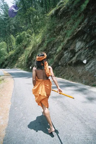 A barefoot man in saffron-orange robes and an orange turban walks away from the camera along a paved mountain road. He carries a long yellow wooden staff in his right hand. The road curves past a steep, grassy rock face on the right and pine trees on the left, with bright sunlight casting a shadow on the asphalt. His back is bare above a wrapped orange lower garment. The figure's identity cannot be determined from appearance, so only the role of walking pilgrim is described.