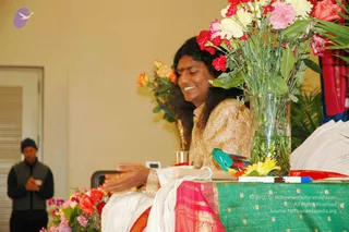 A smiling figure with long dark hair and a red bindi sits behind a table draped in green and pink brocade, with a vase of pink, red and white carnations and roses beside them and a small parrot figurine on the table. The seated person wears a cream and gold embroidered top and has hands raised together near the table. A standing man in a dark knit cap and grey vest with hands folded stands at left in a bright hall with peach walls and an arrangement of orange and pink roses. a person: the seated person has long swept-back hair, a red bindi and a cream embroidered top, and is seated in the honoured position at the convention.