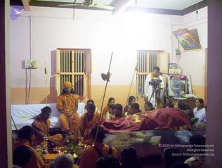 Indoor photo of a final ceremony in a plain room with pink-and-cream walls, a ceiling fan and barred windows. A red-and-gold cloth covers a body lying on a low bed at right. A seated figure at left wears a saffron robe, an orange headwrap and a long beaded necklace; the figure's appearance is uncertain and a candidate identification cannot be confirmed. Around them several people in saffron and white sit on the floor, with brass lamps, a small offering tray and a long staff with a dark fabric piece in front of the bed. A cameraman in white stands on a tripod-mounted camera at the back right. A framed picture hangs on the upper right wall.