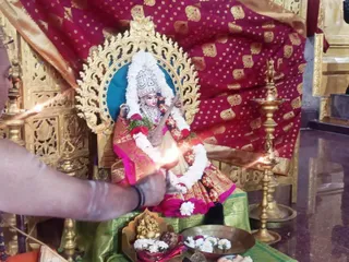 Photo of a temple puja for a Saraswati deity murti seated on a gilded throne with a crown, a veena in its hands, and garlands of white jasmine and red flowers. A priest with bare arms and a wrist thread holds a lit oil lamp or flame toward the deity. Golden oil lamps stand on both sides, a red and gold embroidered cloth backs the throne, and a brass plate with flowers, a small Ganesha figure and offerings sits in front on a green cloth.