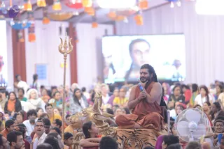 A photograph of a large indoor hall filled with a seated audience. In the centre, a man with long dark hair and a beard, wearing an orange-brown robe, sits cross-legged on a tiger-skin-covered seat in front of a golden bull statue, holding a microphone to his chin. A golden trident stands to the left. A large screen behind shows a close-up of a face, and paper lanterns in orange and blue hang from the ceiling. Many devotees in saris and casual clothes sit on the floor, some with hands raised. a person appearance cues: long swept-back hair, orange ceremonial robe, and a seat set before a golden bull and tiger skin.