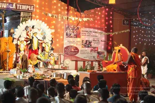 Photograph of an evening puja at a temple hall lit with string lights and warm lamps. A seated figure in saffron and orange robes with a tall orange headpiece sits on a red velvet throne at right. Before him a standing man in orange sannyasi clothing holds a small object and faces the throne. Left, a deity murti with a white floral halo and garlands is decorated with yellow and white flowers, with brass oil lamps, metal vessels and bananas on a cloth-covered table. A banner beside the throne reads Sarva Darshan MEET the Master and lists Satsang, Devotional Worship, Pratyaksha Pada Pooja and Pancha Tapas. Seated devotees fill the foreground, mostly with shaved heads or dark hair.