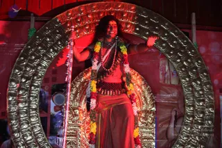 A photograph of a seated figure on an ornate gilded throne framed by a large golden arch with embossed relief patterns, under red stage lighting. The figure wears a saffron-orange cloth draped over the body, a rudraksha mala with multiple strands, a wide beaded waist belt, and a long garland of yellow, orange, red and white flowers over the shoulders. He holds a tall silver staff with ornamental tassels in one hand and rests the other arm on the throne frame. Long dark hair falls to the shoulders and a red mark is on the forehead. Behind the throne are red-draped backdrops and framed photographs. Probable Swamiji (appearance-based label): long swept-back hair, saffron clothing, rudraksha malas and garland cues are visible on a figure seated on a throne.