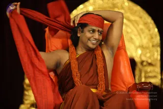 A man seated on an orange-upholstered chair in front of a gold arched backdrop, smiling and raising his arms to adjust a red headband tied over his hair. He wears a rust-red and saffron robe with a yellow-edged waistcloth, a long orange rudraksha-style bead necklace, and a red vibhuti or kumkum dot on his forehead. A long red shawl with printed script hangs from his raised hand. The setting is a stage or dais with a dark curtain.