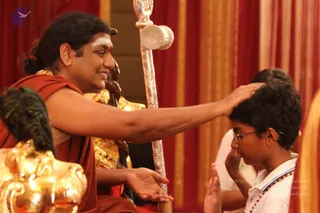 A seated man in saffron-orange robes with a white and red tilak on his forehead and long dark hair tied back, smiling and placing his right hand on the crown of a young boy's head in a blessing gesture. The boy wears glasses, a white shirt and a beaded necklace, and presses his palms together. Behind them stand a silver ceremonial staff and a golden deity murti and red-and-gold drapery. Probable Swamiji cues: long swept-back hair, saffron robes, and ornate gold adornment near the shoulder; he is performing a blessing on a devotee.