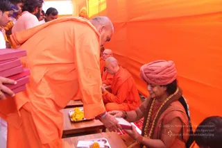 Inside an orange tent, a shaved-headed man in orange robes bends forward at a low wooden table, handing a pink-covered booklet to a woman in a rust-coloured turban, brown beaded necklace and orange blouse with gold bangles, who smiles as she takes it. Behind them, several men in orange robes sit on the floor; one seated monk has a yellow mark on his forehead. Marigold petals sit on a brass tray on the table, and a few people stand at the left edge holding pink boxes.
