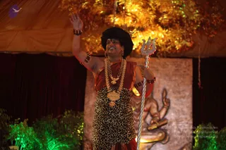 A photograph of a single standing man on a stage under a warm amber canopy with hanging gold lights. He wears a black turban with a small crescent ornament, a red-brown sash over a leopard-print garment, multiple beaded rudraksha-style malas and a heavy gold necklace with pendants, and red-brown bangles. He raises one hand in greeting while holding a silver trishul in the other. Behind him is a carved golden relief of a seated deity murti with a raised hand, and green stage lighting illuminates the foliage at the lower edges.