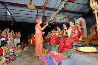 Evening ritual in an open-sided pavilion with a corrugated metal roof and red beams. A barefoot priest in a saffron-orange dhoti with a matching orange head turban stands at centre, bare-chested with a gold-edged cloth draped at the waist, holding a lit oil lamp in one hand and a green leafy branch in the other, offering aarti. Seated on a raised platform at right, two women in red and maroon silk saris wear floral garlands, and a third sits beside them in a dark dress. Along the floor, rows of seated devotees wear garlands and orange cloths, and a man at left holds his hands in anjali. A deity murti sits in a decorated shrine with a trident at the right edge, and a world map hangs on the back wall. Strings of colourful tassels decorate the ceiling.