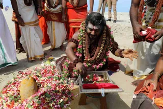 A photograph of an outdoor sandy ritual at a beach or shoreline. A man kneels at the centre, wearing a dark red-brown cloth, a long rudraksha mala, heavy beaded rudraksha necklaces with gold-toned bead ornaments, rudraksha bracelets, and a large garland of pink and red flowers. He has a sandalwood or vibhuti mark on his forehead and tends a wooden tray lined with red velvet holding flower petals and garlands. Beside him lies a large heap of scattered flowers, pink, white and red petals, and a yellow-ochre conical stone. Around them, several men stand in white dhotis with gold borders and saffron-orange cloths, with rudraksha malas, and one holds a red cloth. A uniformed police officer stands in the background.