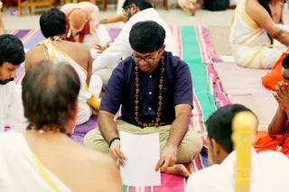 Indoor photograph of a seated group on colourful striped mats, with devotees in white dhotis and yellow-bordered shawls and some in orange robes. In the centre a young man in a dark navy shirt, khaki trousers, glasses and a long rudraksha mala holds a sheet of white paper and reads from it. Around him people sit cross-legged, one man in the foreground shows a bare back and shaved head, and a yellow-handled object appears at the lower right. No figure is identified as a person by appearance cues here.