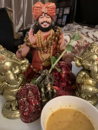 A photograph of a home altar arrangement. A cutout or printed image of a smiling bearded man in an orange-red turban with a gold emblem, wearing layered gold and rudraksha garlands, raises one hand in blessing; this is a depiction on a flat cutout, not a physical person, and the appearance cues are a turban, saffron clothing and rudraksha malas, so it is a probable depiction of Swamiji. Around it stand two gold-painted Ganesha figures, a small dark bronze Ganesha, and a red carved Ganesha plaque and figurine. A green leafy sprig lies across the arrangement, and a white bowl holds a yellow liquid in the foreground. A bed with floral bedding is visible behind.