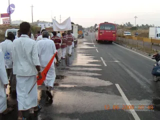 A road procession on a wet highway at dusk. Several men in white dhotis and white shirts walk along the left lane, one carrying an orange flag on a pole. Behind them, men in maroon and red uniforms with gold trim walk beside a white cloth banner held aloft. A red state bus travels in the right lane, and a white truck and motorbikes follow. Roadside signboards and power lines line the background.