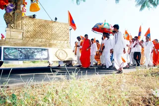 Outdoor procession on a paved road beside dry grass. A golden ratha-style chariot with carved panels and a painted banner stands at left. Walking alongside it are devotees in white dhotis and white shirts wearing rudraksha malas, and a central figure in a saffron-orange robe. Several people carry saffron and orange flags, and a multicoloured umbrella is held over the central figure. The central figure's face and identity are not established by visible cues here; he is described by role only as a robed participant. Several women in orange saris walk at right. The scene is a street procession.