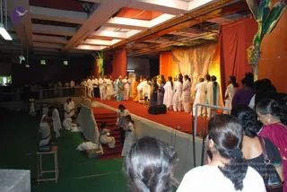 Interior hall photographed from the side during a ceremony. A raised red-carpeted stage with orange and red draped backdrop, a banana-plant decoration, and a painted gold-and-cream panel stands at the far end. Several people in white garments stand on the stage, including a figure in an orange-saffron garment and women in saffron and white saris. Below, on the green floor, attendants in white kneel and sit near a low wooden table and boxes, apparently arranging items. In the foreground, women with hair in buns and a woman in a magenta sari watch from the crowd. A monitor speaker stands at the stage edge. No identifiable a person depiction is clearly visible; the orange-clad figure's appearance is not distinctive enough to confirm.