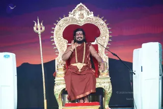 A man with long dark hair and a beard sits on an ornate gold throne with a red velvet back and a gold crest at its top. He wears a red-orange saffron robe draped over the shoulder, a gold necklace of beaded strands and a large gold pendant at the chest, gold bangles on both wrists, and a red bindi. He raises one hand in a blessing gesture while smiling, with a microphone stand at his side. A golden trident stands to the left, and a dusky red and purple sunset backdrop with a dark mountain silhouette fills the stage behind him. White air-conditioning units flank the stage.